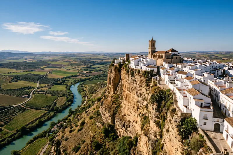 Pueblo blanco asomado al borde de un tajo rocoso, sobre un río y campos de olivar de la provincia de Cádiz